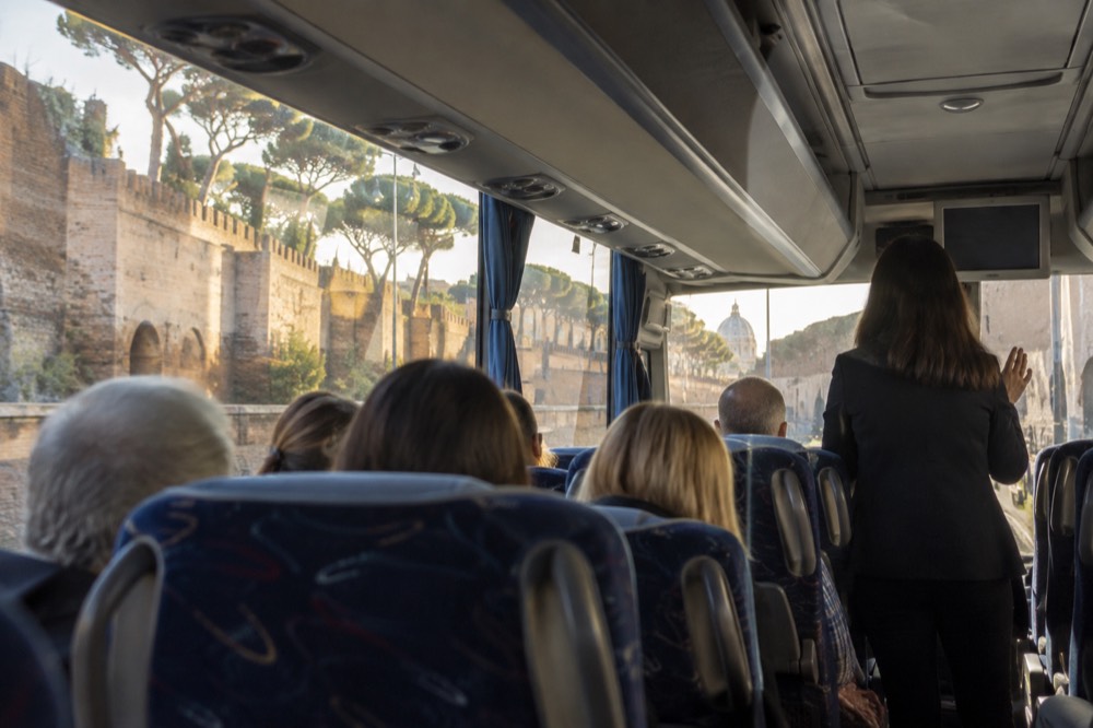 A tour director holding a sign and speaking to a group of seated passengers inside a coach, with a mix of ages visible