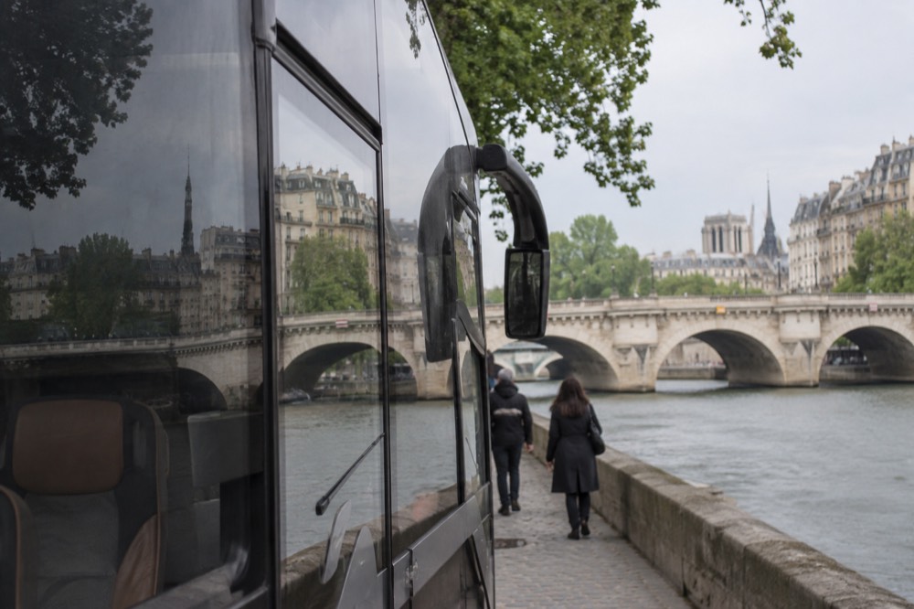 A touring coach parked beside a stone bridge over a river in an old European town