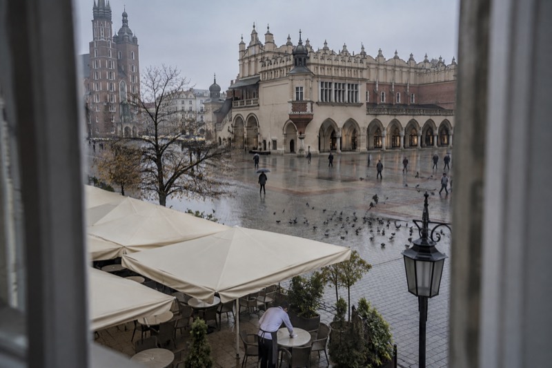 Grey November morning over the Rynek Glowny in Krakow, Poland, wet cobblestones around the pale arcaded Cloth Hall, pigeons scattering and a waiter wiping a table beneath cloth awnings.