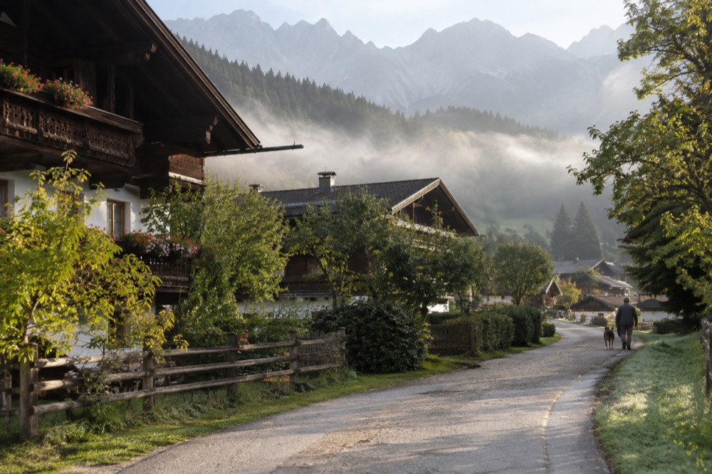 A country road winding past alpine houses toward distant mountains