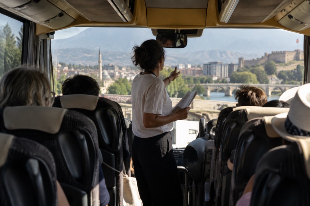 A tour director standing at the front of a coach aisle speaking toward the seats
